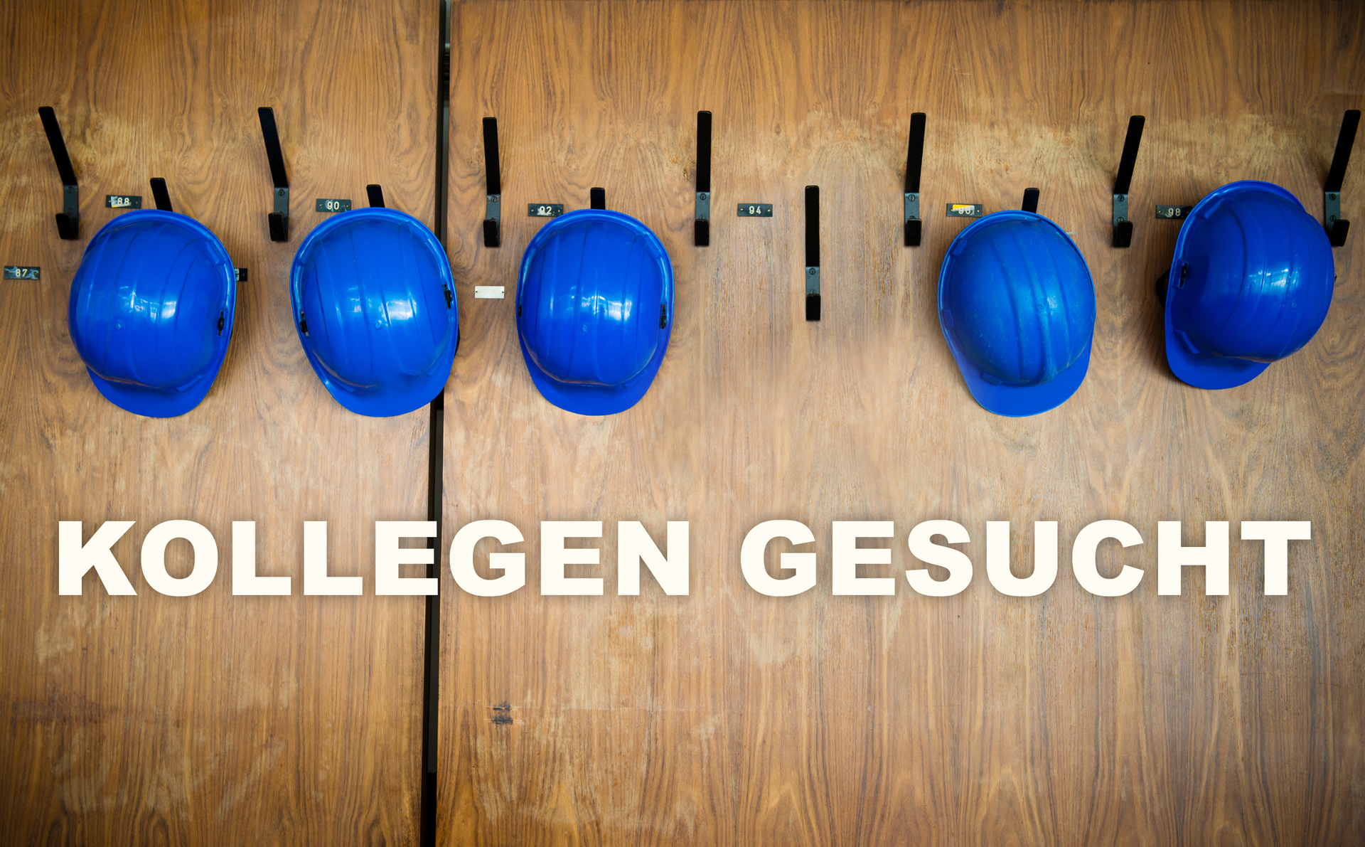 An einer Holzwand in einem Bauwagen hängen 5 blaue Bauhelme - ein Haken ist noch frei